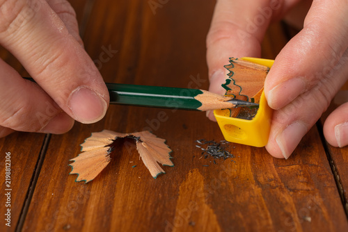 Close-up of human hands sharpening a green wooden pencil with a yellow manual sharpener, showing wooden shavings on a wooden table.