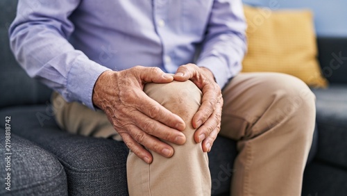 Senior man holding painful knee while sitting on a sofa indoors
