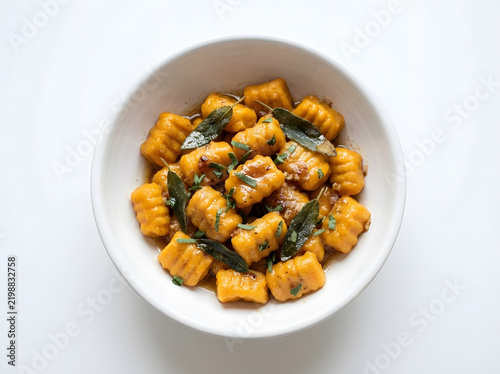 An overhead view of delicious homemade pumpkin gnocchi served with aromatic fried sage leaves and rich melted brown butter sauce in a clean white ceramic bowl over a bright white background