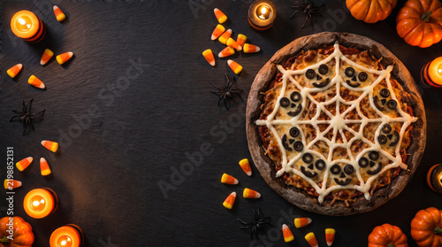 Halloween pizza flat lay with spider web cheese, olive spider, pumpkin, candy corn, candle, and black background creating spooky mood