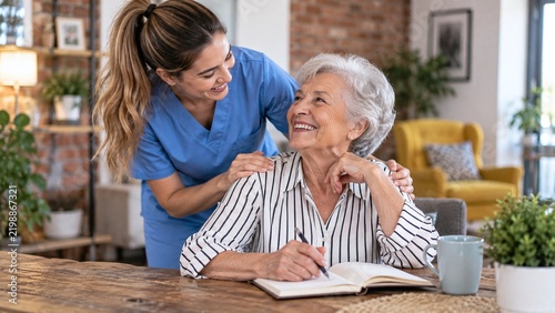 Caregiver providing compassionate support to a happy senior woman writing in a notebook at home, illustrating elderly care and companionship