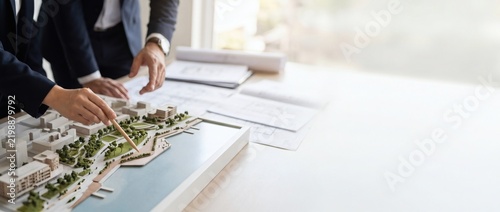 Architectural team hands examining detailed physical scale model of urban masterplan on wooden table. Architects collaborating on sustainable city building construction schematics in studio.