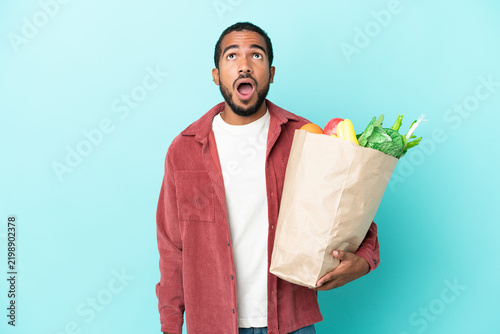 Young latin man holding a grocery shopping bag isolated on blue background looking up and with surprised expression