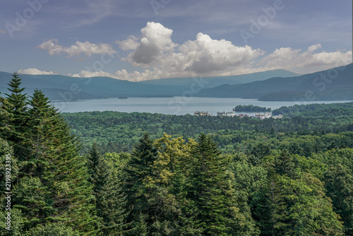 Panoramic view of Lake Akan in Hokkaido, Japan, surrounded by dense forests and mountain ranges beneath a blue summer sky. Lake Akan is part of Akan-Mashu National Park in eastern Hokkaido.