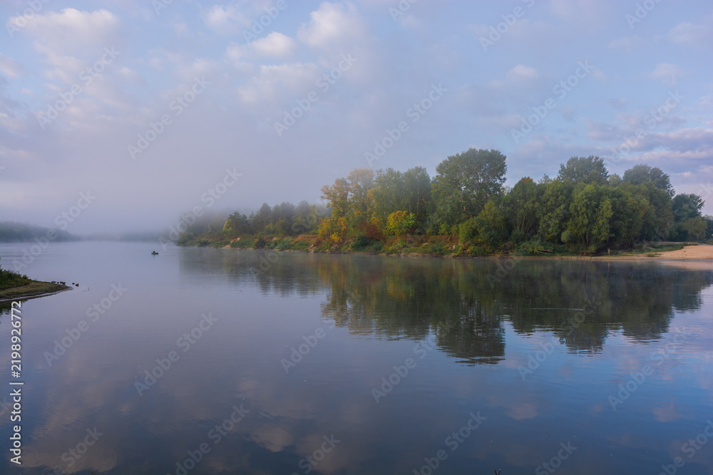 Obraz premium A peaceful river flows calmly under a cloudy sky, reflecting the green trees lined along the misty distant shoreline
