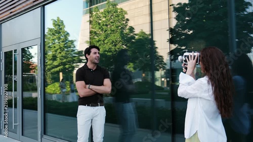 Woman filming a handsome man posing in front of a modern glass building with a retro camera