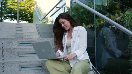 Beautiful woman sitting on modern stairs, typing on her computer and smiling happily