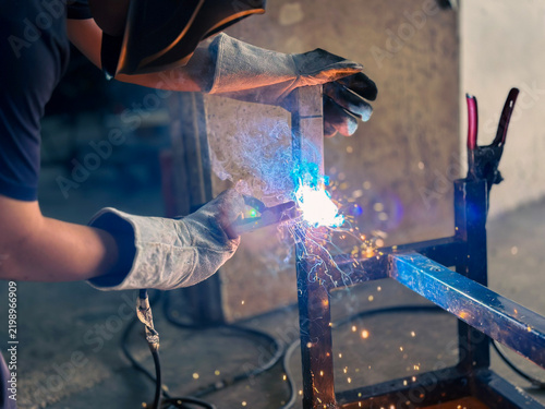 Welder using arc welding torch with bright sparks and smoke on metal frame in industrial factory workshop.