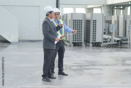 Rooftop HVAC inspection engineer manager hard hat safety vest clipboard, two male professional review maintenance plan near air conditioning unit on commercial building roof, focused teamwork
