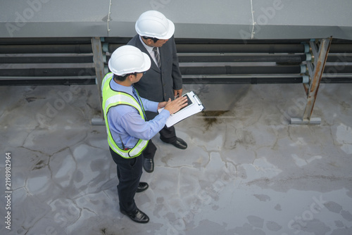 Engineer inspector hardhat clipboard safety vest rooftop industrial inspection maintenance pipe concrete teamwork, two men review facility condition with caution and focus