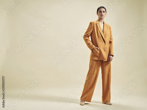 A woman posing in a mustard yellow oversized blazer and matching wide leg trousers with a hand in her pocket against a neutral studio backdrop