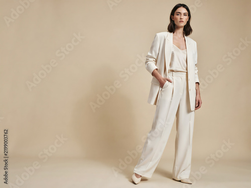 A woman standing in a full length shot wearing a cream colored oversized blazer and matching wide leg trousers against a neutral background