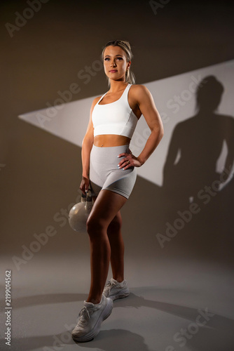 Fit young woman in white workout gear holding kettlebell in studio with dramatic lighting shadow