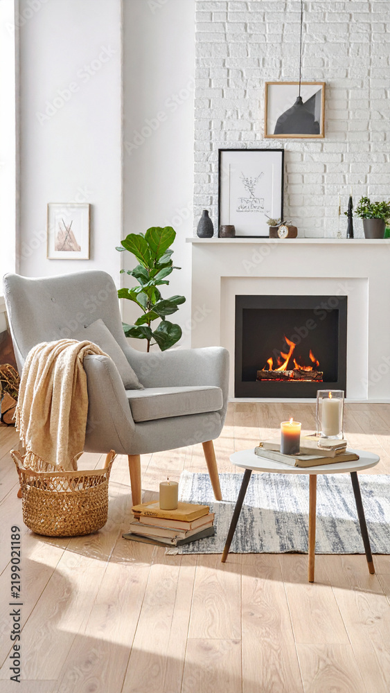 Obraz premium A cozy living room interior with a warm fireplace, grey armchair, and soft decor. This peaceful space offers a serene, inviting atmosphere, perfect for reading. Nobody, no people present.