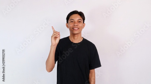 Bild auf Leinwand Young Asian man in a black t-shirt smiling and pointing his index finger upward against a white studio background