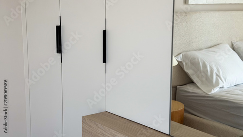 Modern neutral bedroom interior with built-in white wardrobe, large mirror, wooden furniture and cozy bed, contemporary minimalist home design.