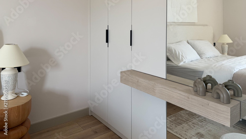 Modern neutral bedroom interior with built-in white wardrobe, large mirror, wooden furniture and cozy bed, contemporary minimalist home design.