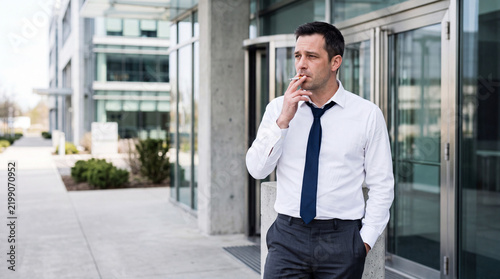 Businessman smoking a cigarette outside a modern office building. Stressed male employee taking a smoke break during workday. Unhealthy habit and corporate stress concept