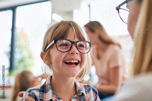 Young girl with blonde hair and oversized dark framed glasses laughing brightly while interacting with an ophthalmologist in a brightly lit clinic, mother blurred in background near large windows