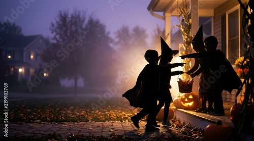 Cinematic silhouettes of children in costumes trick-or-treating on a misty porch with glowing pumpkins and wide copy space for Halloween banner.