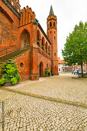 Side view of a historic Gothic brick town hall with exterior staircase, arched details and slender tower, photographed in portrait format.