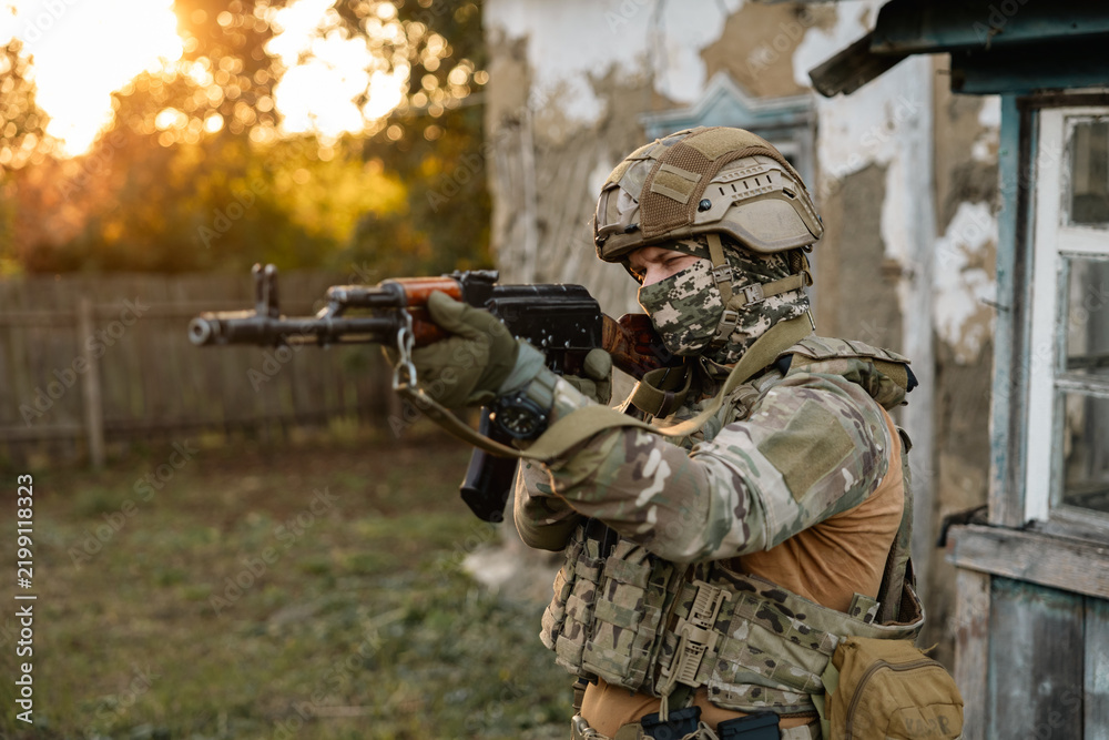 Fototapeta premium Military soldier posing at camera with assault rifle against the backdrop of an old village house