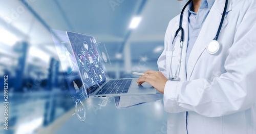 A doctor in a white lab coat with a stethoscope holding and using a laptop displaying medical data and technology graphics.