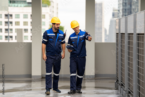 Industrial engineer and technician inspect rooftop equipment beside commercial building ventilation units, wearing protective helmet and work uniform during professional maintenance