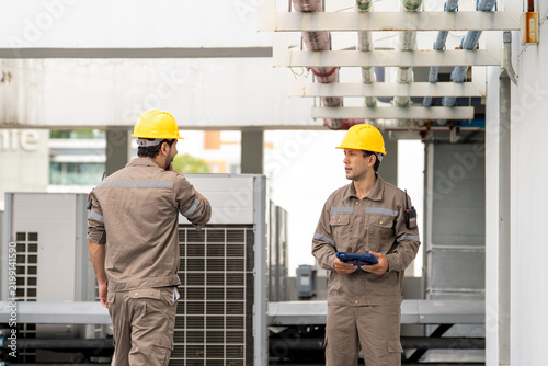 Engineer industrial worker wearing yellow helmet and uniform during equipment inspection, holding tablet inside modern power facility with serious focus and teamwork