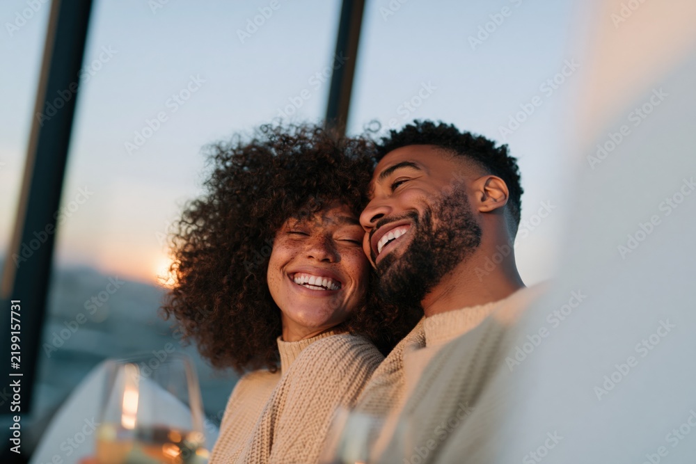 Fototapeta premium Smiling couple embracing on balcony at sunset with wine glasses for romantic lifestyle banner and background design