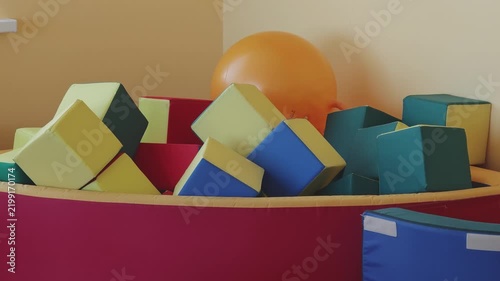 Brightly colored soft foam blocks filling a ball pit in a children's playroom. A large inflatable ball rests in the corner of the room
