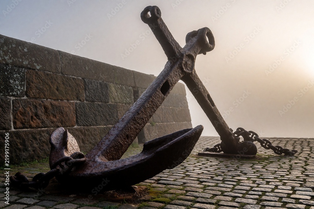 Fototapeta premium Old rusty anchors on cobblestone quay in morning fog