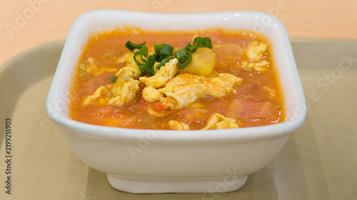 Close-up of Chinese style tomato and scrambled egg soup served in a white bowl, garnished with chopped spring onions and featuring a savory tomato broth.