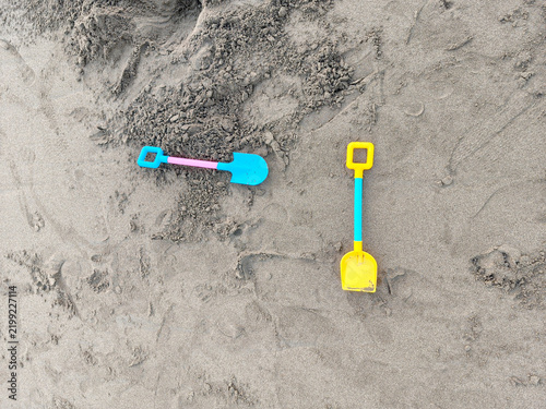 Two colorful plastic toy shovels lay on the beach sand. One shovel was yellow with a blue handle, while the other was blue with a pink handle.