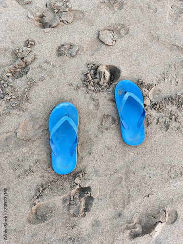 A pair of bright blue sandals rests on the textured beach sand, surrounded by a few footprints, capturing a simple, relaxed moment by the shore.