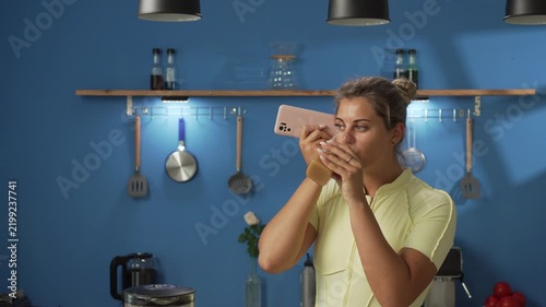 woman in sportswear listening voice message or voicemail on phone and drinking smoothie in the kitchen. Technology and healthy lifestyle concept