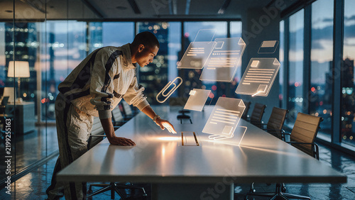 Man interacts with glowing holographic document icons hovering above a long conference table in a high-rise office at dusk. Warm orange light emanates from the digital interface.