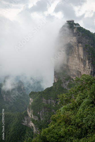 Fotografie A dramatic vertical drop of a towering rock pillar at Tianmen Mountain, featuring a high-altitude observation deck emerging through a thick layer of morning mist
