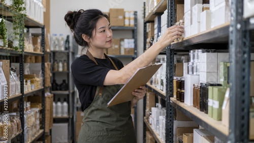 Committed Asian Small Business Owner Methodically Checking Stock Levels with Clipboard in Backroom Storage Area Filled with Neatly Arranged Products, Showcasing Practical Retail Operations.
