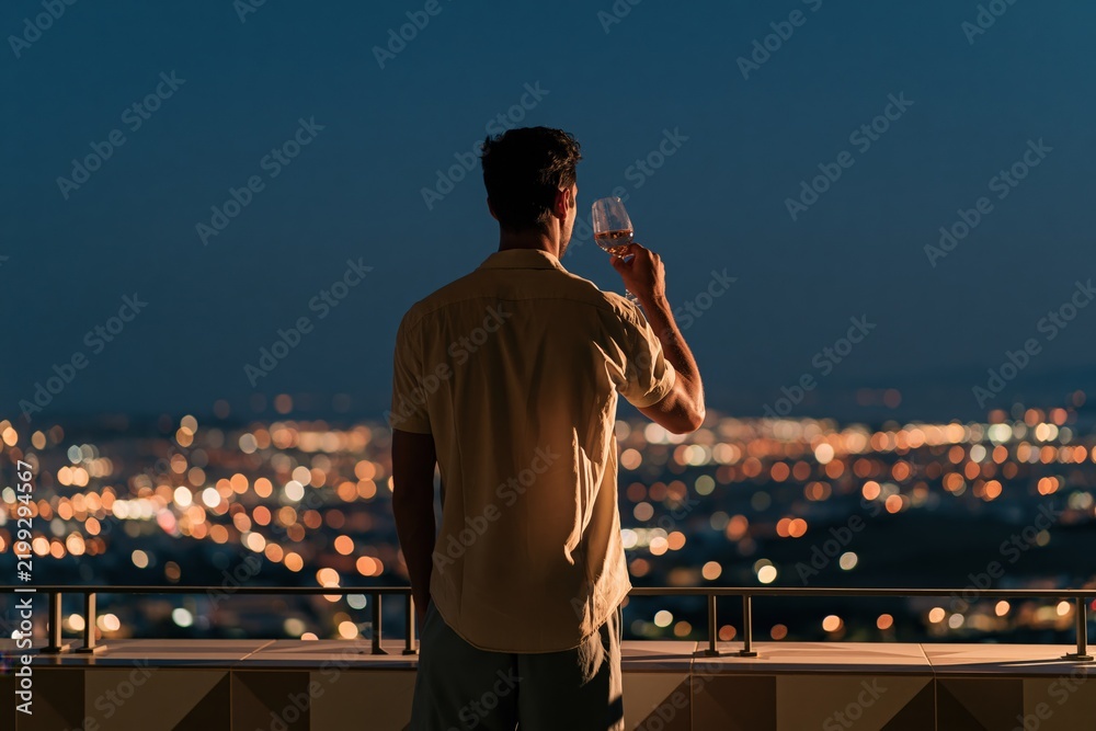 Fototapeta premium Man holding wine glass on balcony overlooking city night skyline with bokeh lights for rooftop lifestyle background and evening relaxation banner