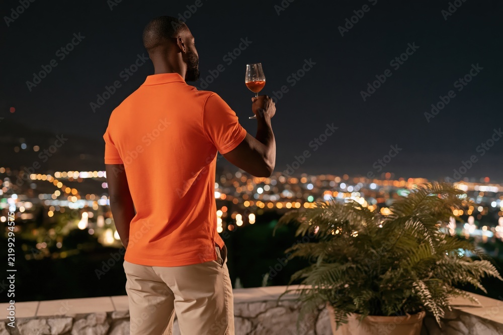 Fototapeta premium Man in orange polo raising a wine glass on a terrace overlooking the city night skyline for lifestyle poster and rooftop background design