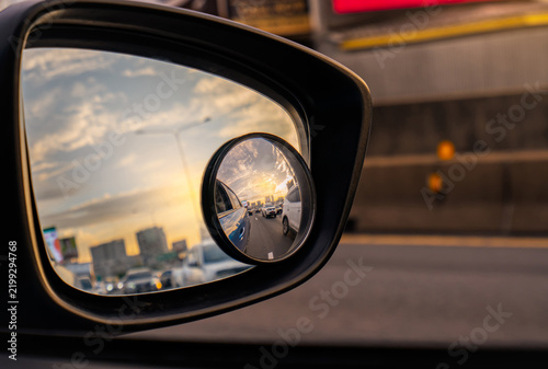 Car side mirror reflecting traffic on urban road at sunset, with convex blind spot mirror showing nearby vehicles for safer driving, road awareness and transportation safety. Accident prevention.