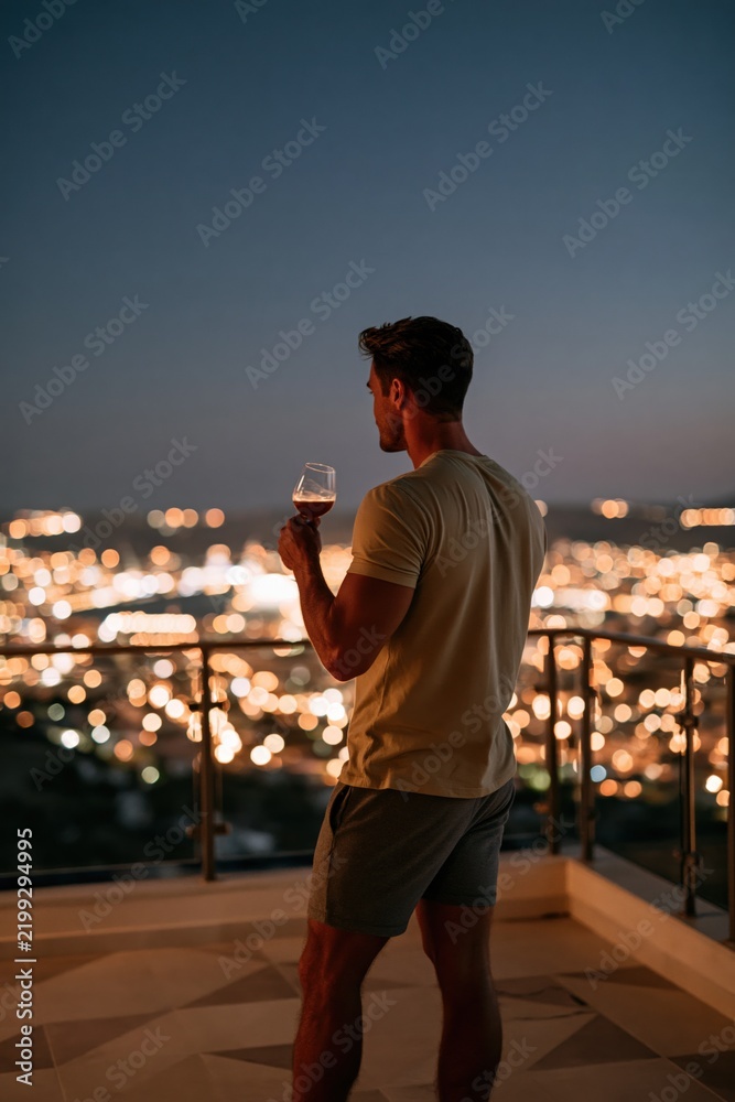 Fototapeta premium Man holding wine glass on balcony at night overlooking city lights for lifestyle poster and background