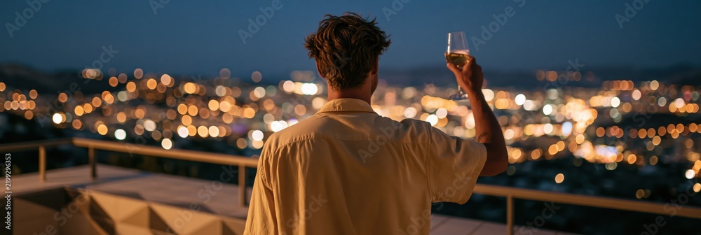 Obraz premium Back view of a man raising a wine glass on a rooftop with city bokeh lights background for banner and wallpaper design