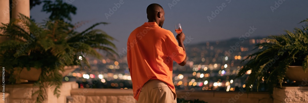 Obraz premium Man on terrace holding a glass while overlooking city lights at dusk for evening banner and travel lifestyle background