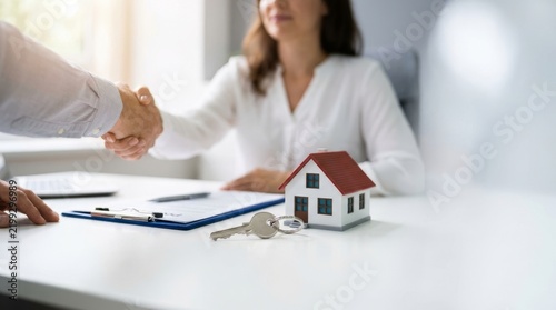 Two people shaking hands across a desk with a house model and keys, symbolizing a real estate agreement in an office setting.