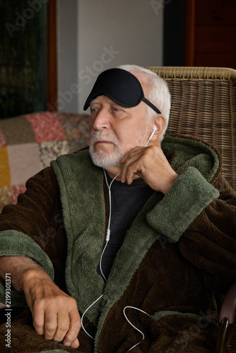 Картината върху платно Elderly man resting in a cozy chair wearing an eye mask and listening to music a