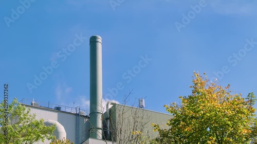 Industrial facility with a tall chimney and visible clean steam rising from the roof, surrounded by autumn trees under a clear blue sky, clean industrial technology concept