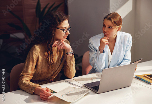 Two caucasian women rest their chins on their hands, facing each other across a sunlit white desk, where printed line charts, a notebook and a laptop lie between them.