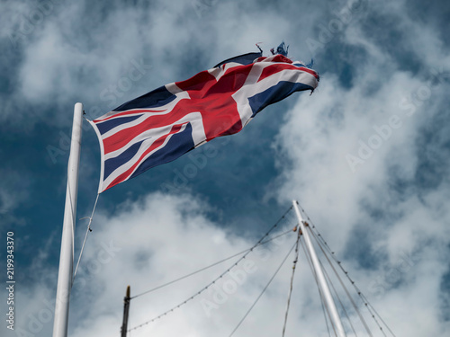 The Union Jack lifts diagonally from the mast of a sailing ship, its flying edge worn thin against blue sky and white clouds.
